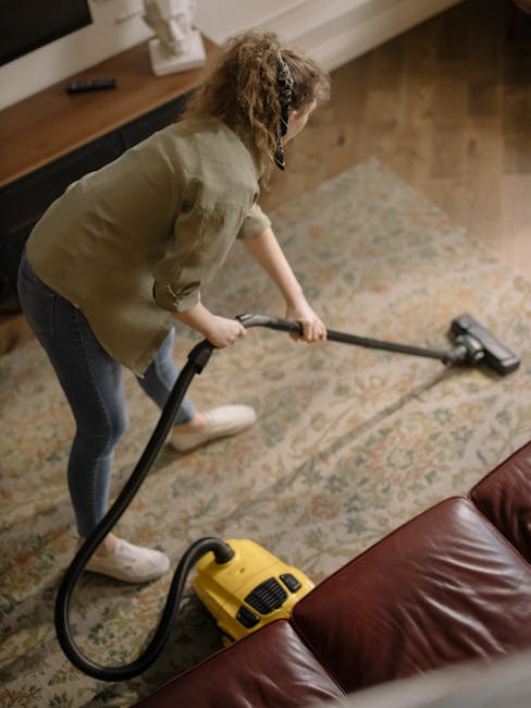 A professional cleaner dressed in a white protective suit, gloves, and blue shoe covers is performing deep carpet cleaning in a bright, modern living room. The cleaner is using a vacuum or steam clean