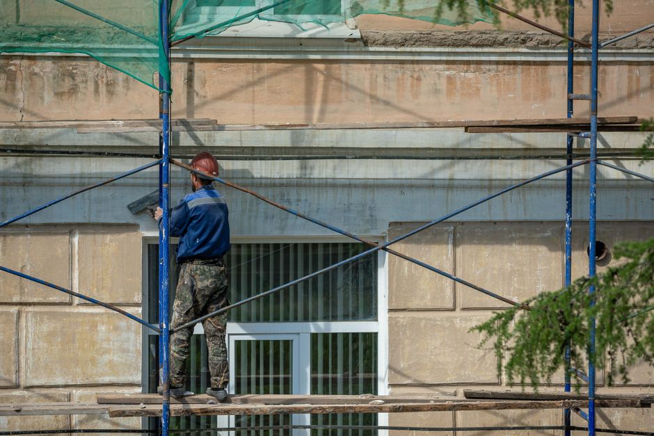 A construction worker wearing a red safety helmet and camouflage trousers stands on a wooden plank platform within blue scaffolding erected around the exterior of a building. The worker is engaged in 