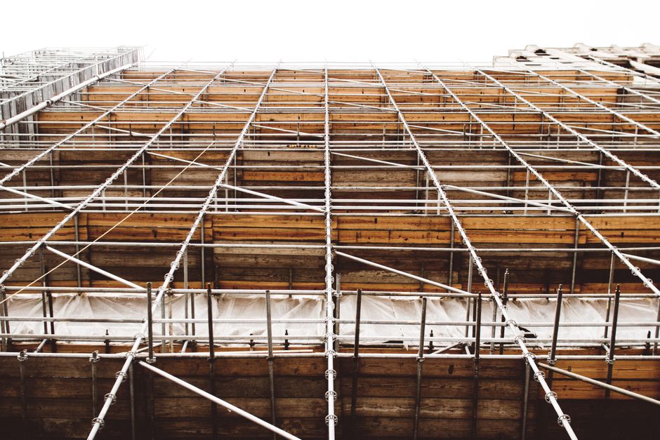 A construction worker wearing a red safety helmet and camouflage trousers stands on a wooden plank platform within blue scaffolding erected around the exterior of a building. The worker is engaged in 