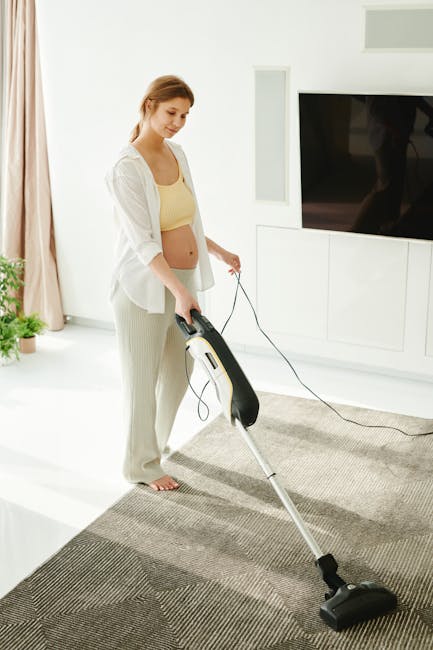 A young woman with light brown hair, dressed in a pale yellow top and light-colored trousers, is vacuuming a modern living room with a handheld vacuum cleaner. The room features a white wall mounted with a large flat-screen TV and built-in cabinets beneath it, with a light-colored wooden floor partially covered by a patterned area rug. The space is well-lit with natural sunlight coming through a window, and a potted plant is visible in the background. The woman is engaged in surface cleaning, focusing on the area rug, and Harringay Cleaners offers professional domestic cleaning services, including deep cleaning and sanitisation, ensuring surfaces such as carpets and flooring are free of dust and dirt in residential spaces.