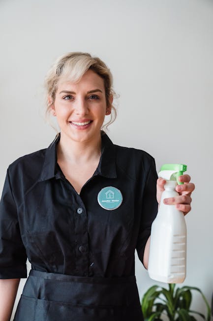 A smiling woman with blonde hair wearing a black uniform and a name badge stands indoors, holding a white spray bottle with a green cap, preparing for cleaning tasks. The background features a plain, light-colored wall and a green potted plant, indicating a clean and organized environment suitable for surface cleaning, deep cleaning, or sanitisation. The image highlights professional cleaning practices by Harringay Cleaners, emphasizing their commitment to hygiene and thorough cleaning services in residential or commercial settings.